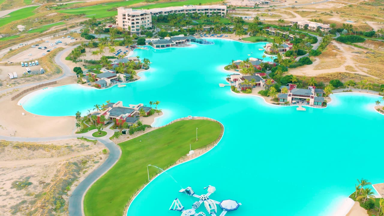 Drone view of turquoise lagoon, curved road, lawn, sandy lots, and resort buildings.