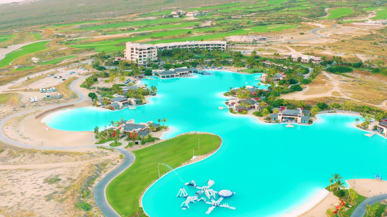 Aerial frame of Diamante Cabo San Lucas showing a turquoise lagoon, curved sandy beach, green lawn, small buildings, roads, parked vehicles, and inland fields at the start of the clip.