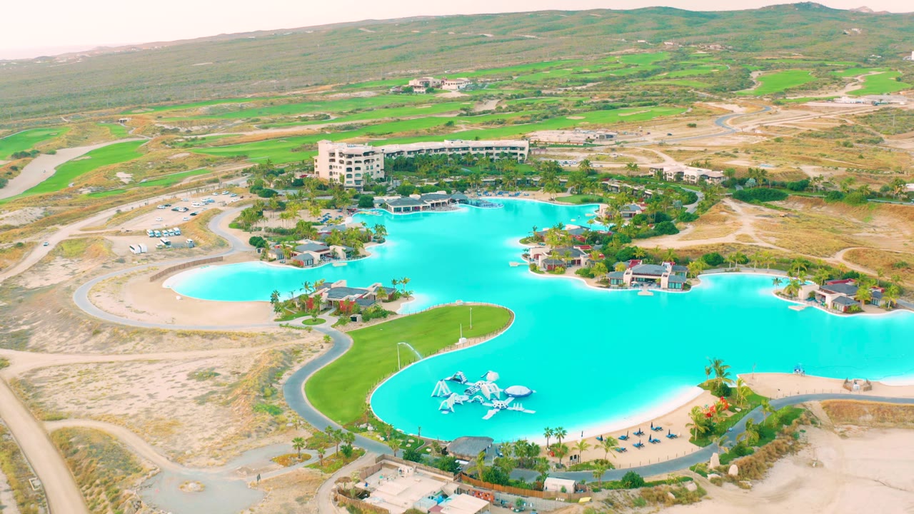 Aerial frame over Diamante Cabo San Lucas showing the blue lagoon shifted slightly in view, with a pale sandy basin at left, curving roads, green banks, buildings, and no visible people.