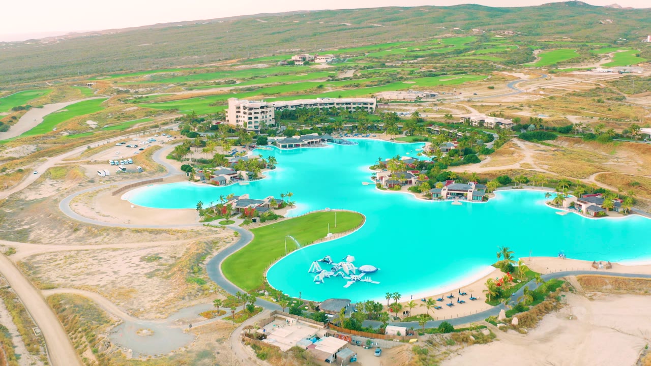 Aerial view in Diamante Cabo San Lucas with the turquoise water feature across the lower center, a small green peninsula, surrounding dry lots, low buildings, and no visible people.