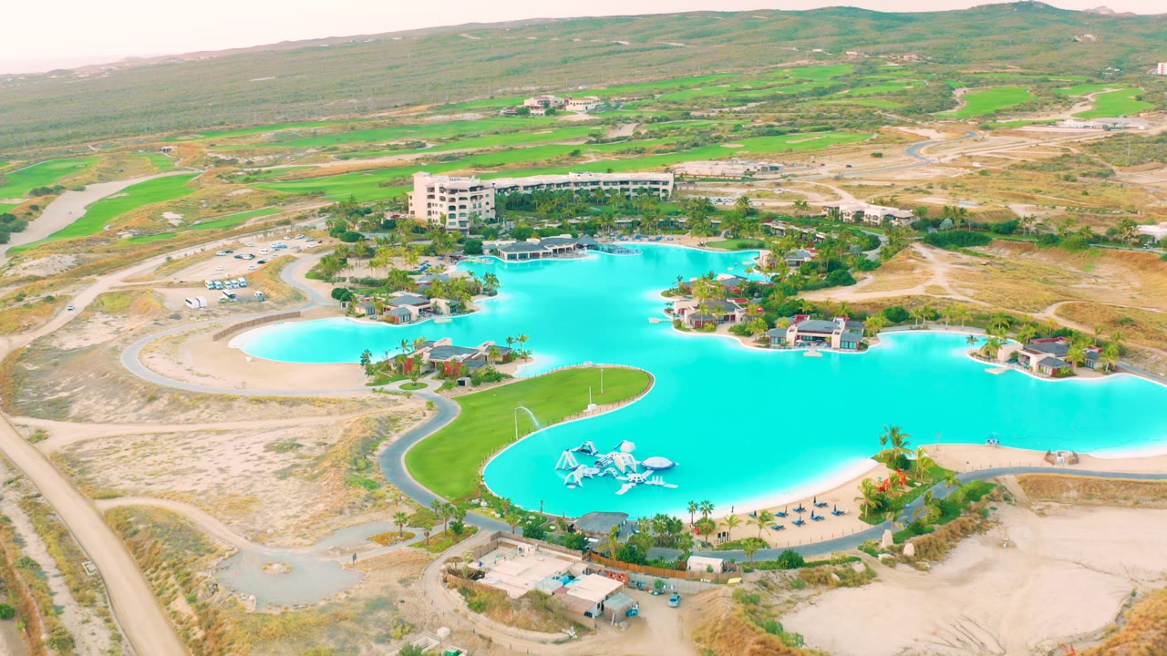 Mid-clip aerial frame over Diamante Cabo San Lucas showing the lagoon's broad right edge, a green lawn strip in front, sandy roads and dry ground around it, buildings beyond, and no visible people.