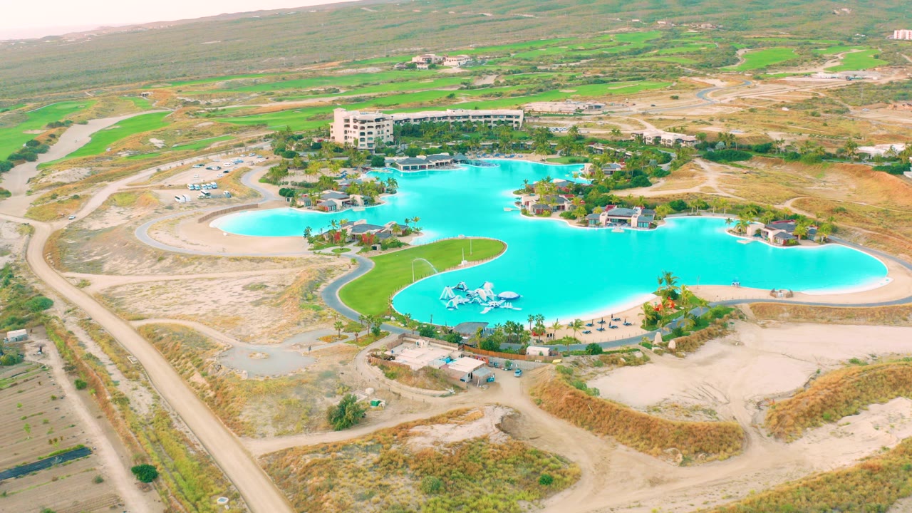 Early aerial frame over Diamante Cabo San Lucas showing a large turquoise pool, curved dirt roads, landscaped lawn patches, a nearby building, and no visible people.