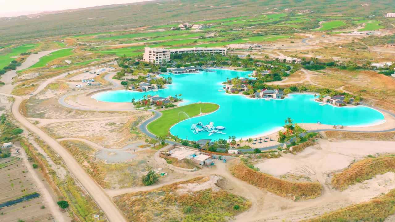 Aerial view in Diamante Cabo San Lucas with the turquoise pool centered, a pale sandy basin to the left, dirt access roads, green fields beyond, and no visible people.