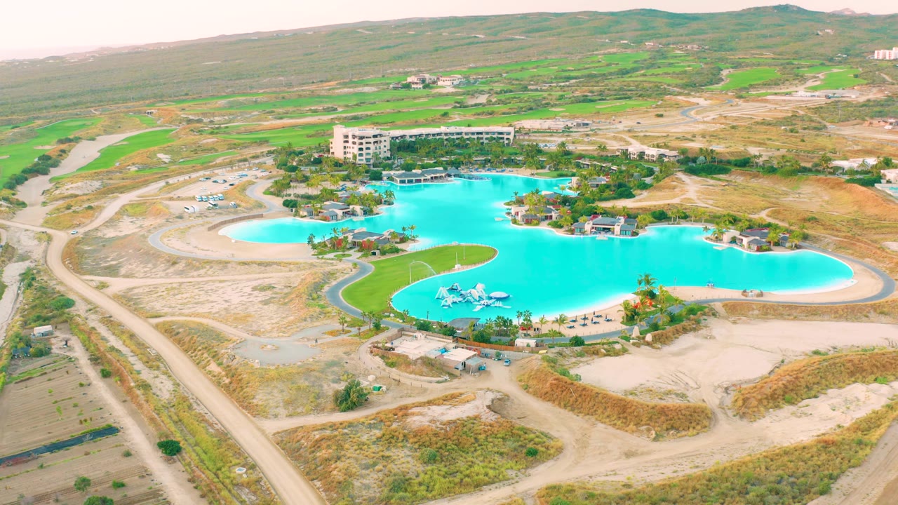 Wide aerial frame in Diamante Cabo San Lucas with the turquoise pool across the center, sandy lots in front, a road corridor at left, a building behind the water, and no visible people.