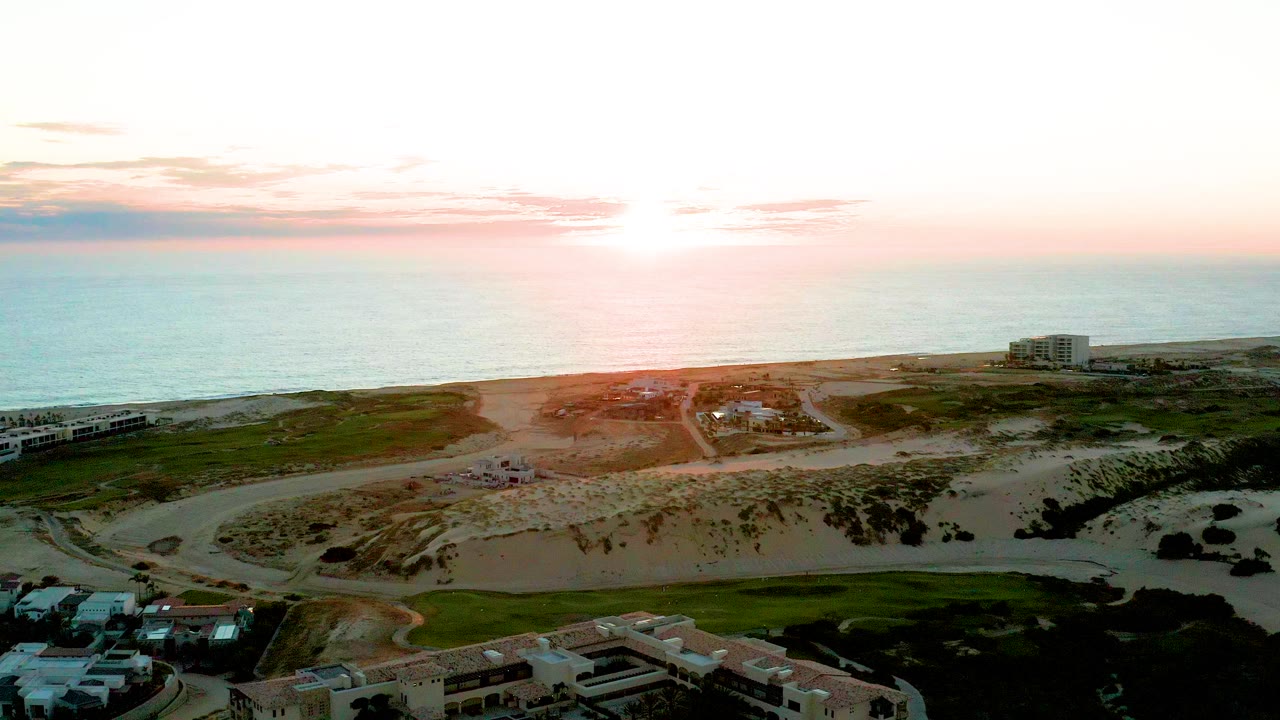 Opening aerial frame over Diamante Cabo San Lucas shows the ocean horizon, sandy dunes, coastal roads, scattered residential buildings, green grass, and no visible people.