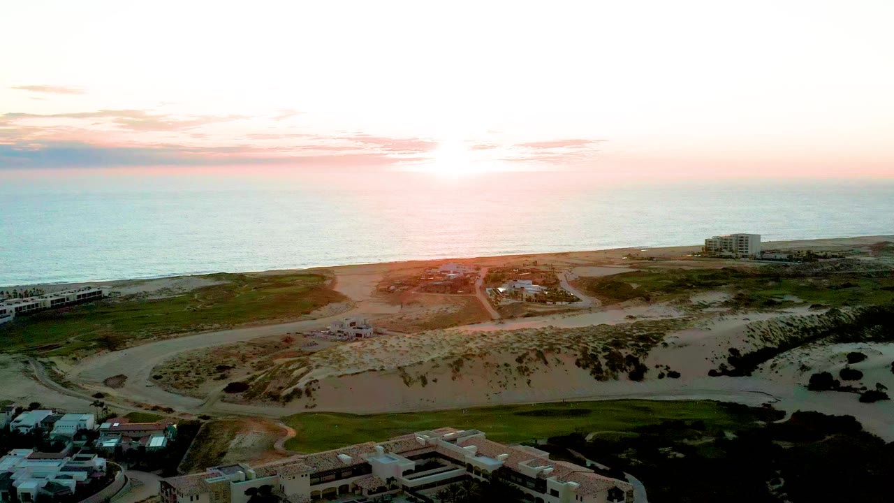 At 0:01, the Diamante Cabo San Lucas aerial view holds on the beach, dunes, roadway curves, residential structures, soft sunset sky, and no visible people.