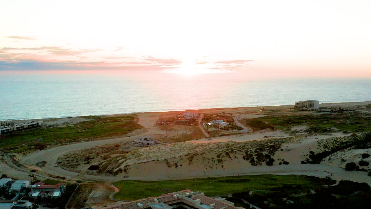 At 0:00, an aerial view over Diamante Cabo San Lucas shows the sun above the ocean horizon with sandy dunes, a curving road, green patches, coastal buildings, and no visible people.