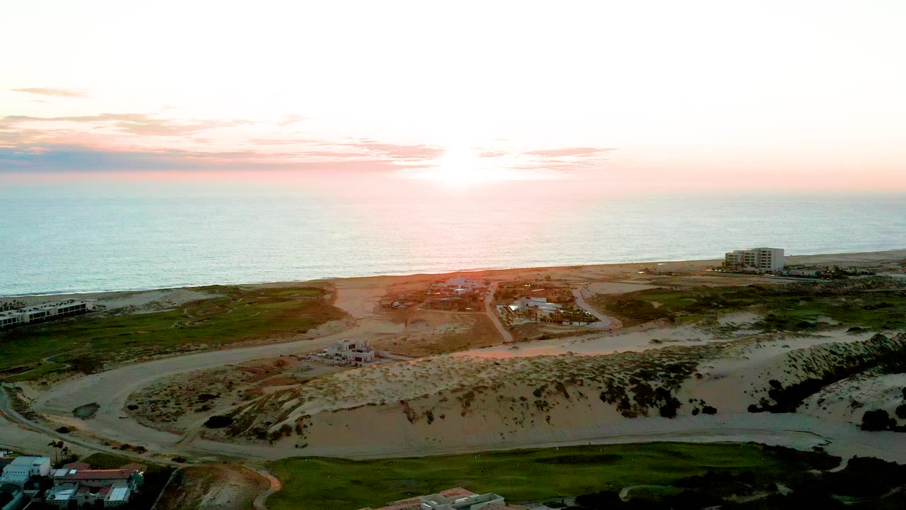 Bright sunset above ocean with dunes, beach road, grass, and buildings.