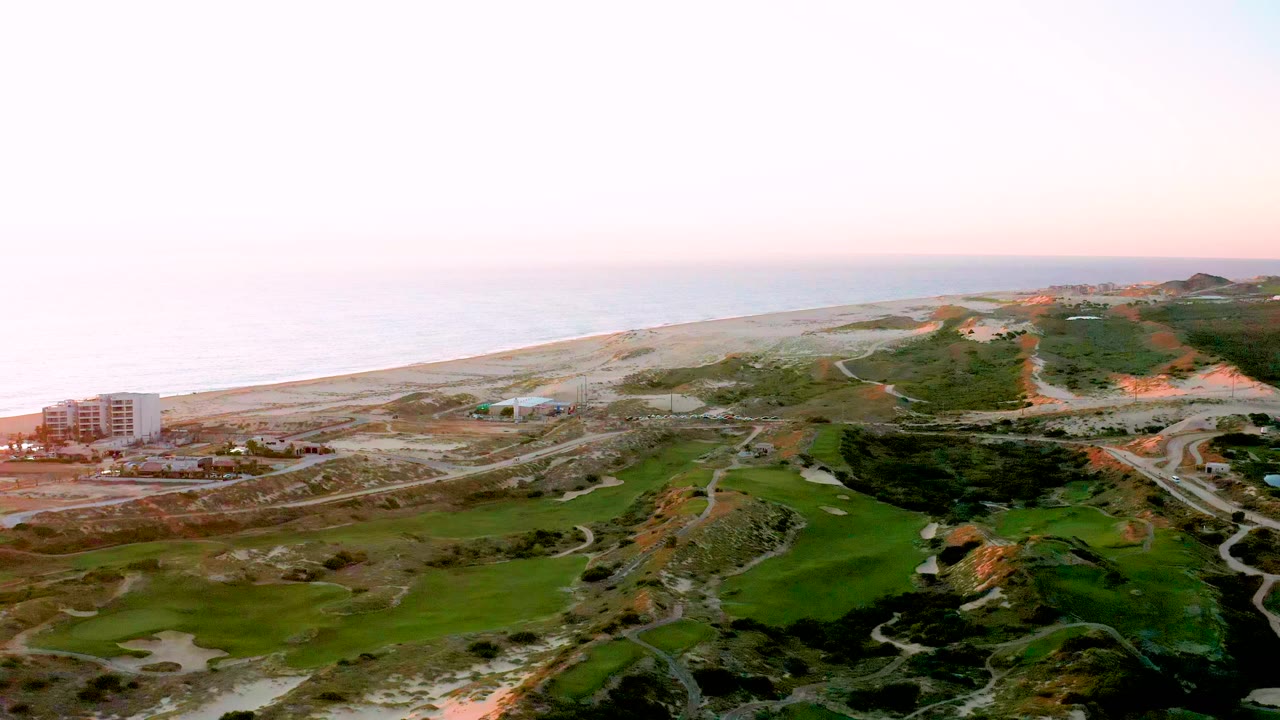 Aerial view in Diamante Cabo San Lucas showing a pale ocean horizon, sandy beach band, green landscaped terrain, winding roads, a left-side building near the shore, and no visible people.