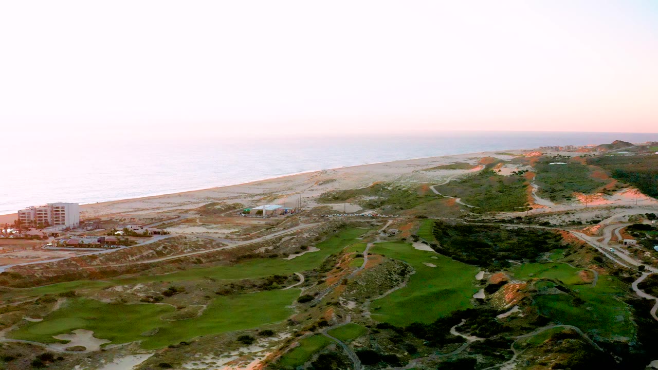 Wide aerial frame over Diamante Cabo San Lucas with the ocean and beach stretching across the background, a building near the left edge, green terrain below, sandy hills to the right, and no visible people.