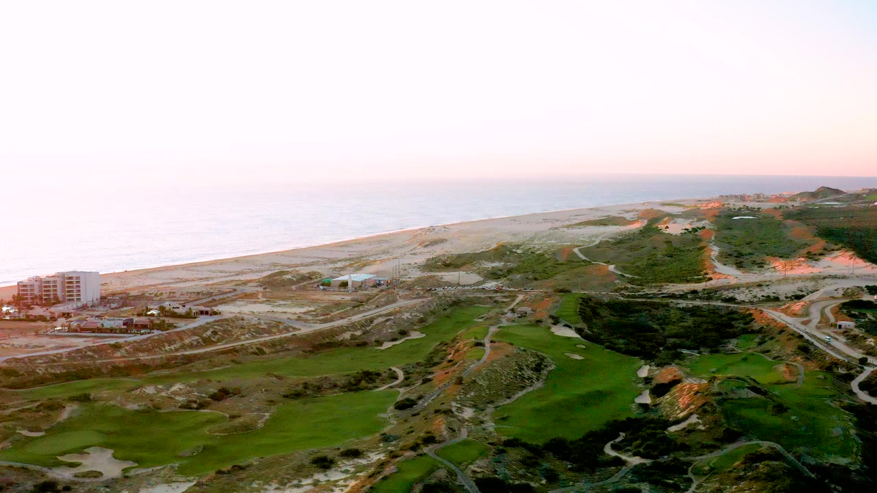 Aerial coastal scene with beach, ocean, building, roads, and green fairway-like land.