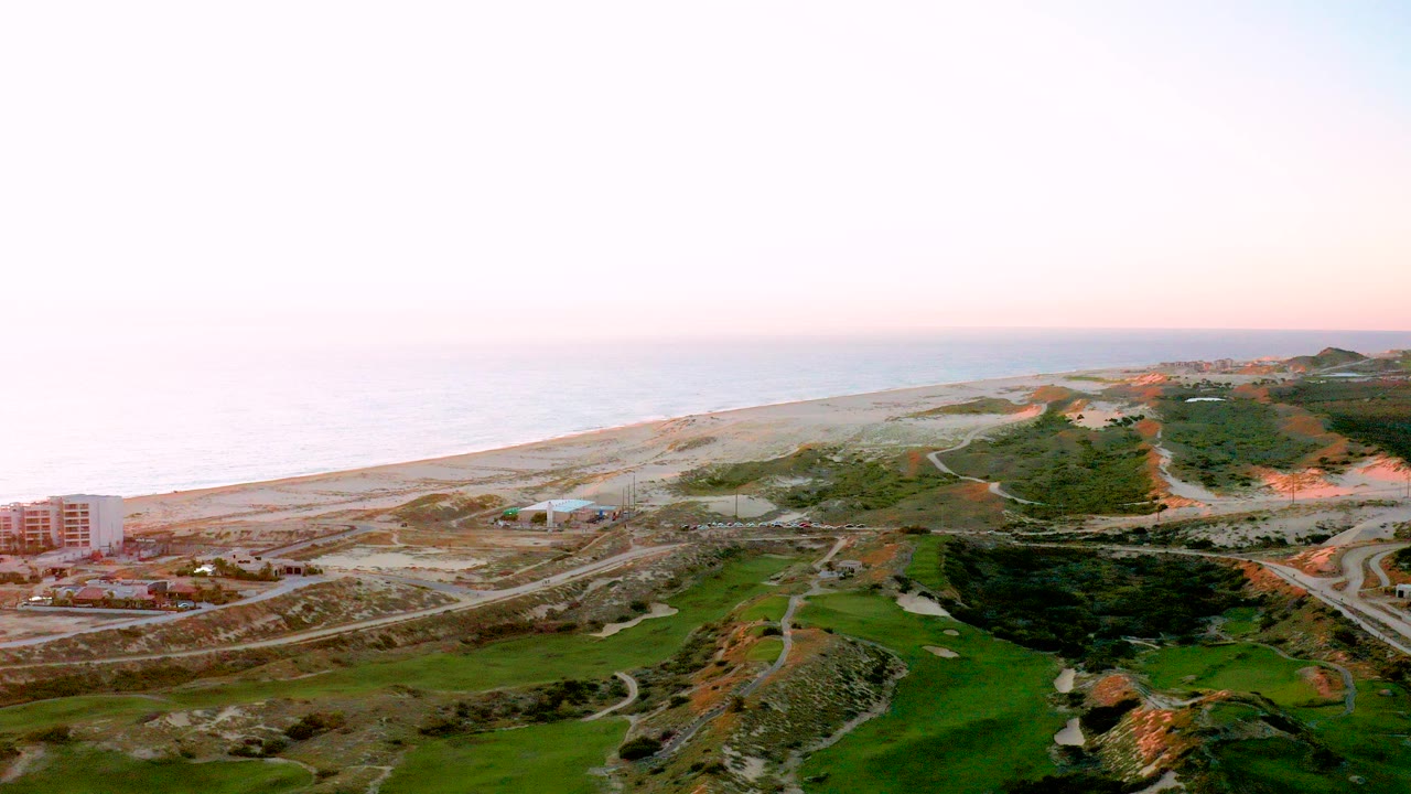 Aerial view of ocean, beach, roads, green terrain, and shoreline buildings at sunset.