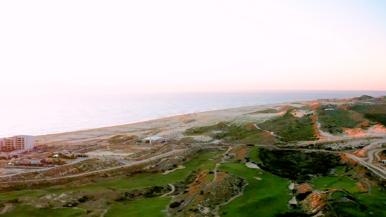Aerial frame of Diamante Cabo San Lucas showing the pale beach and ocean horizon, a road network through sandy development, green foreground terrain, buildings near the coast, and no visible people.