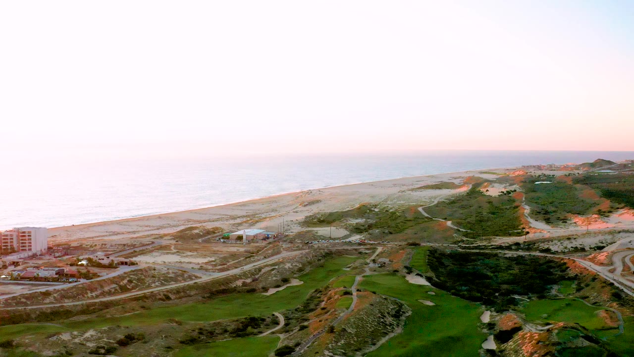 Aerial sunset view over Diamante Cabo San Lucas with the ocean and beach stretching left to right, developed roads and structures in the midground, darker green terrain below, and no visible people.