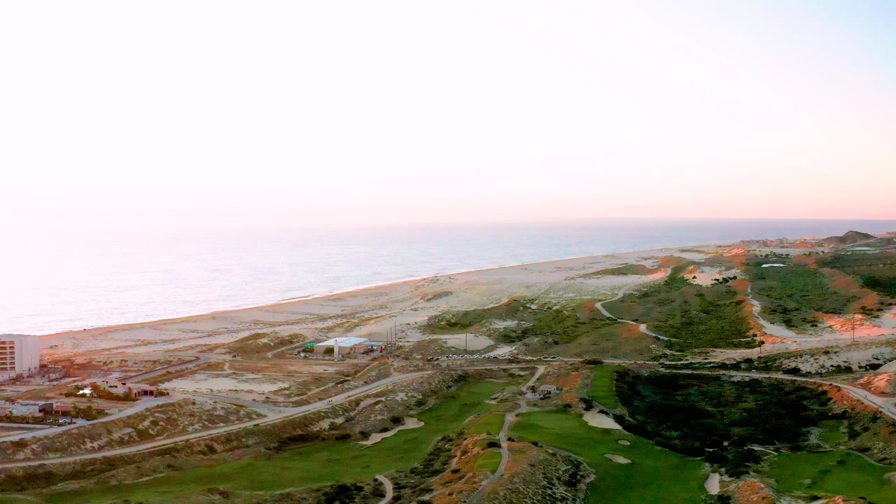 A second-wide view of Diamante Cabo San Lucas shows the ocean horizon, pale sandy shoreline, winding road with vehicles, green fairway-like terrain, and no visible people.