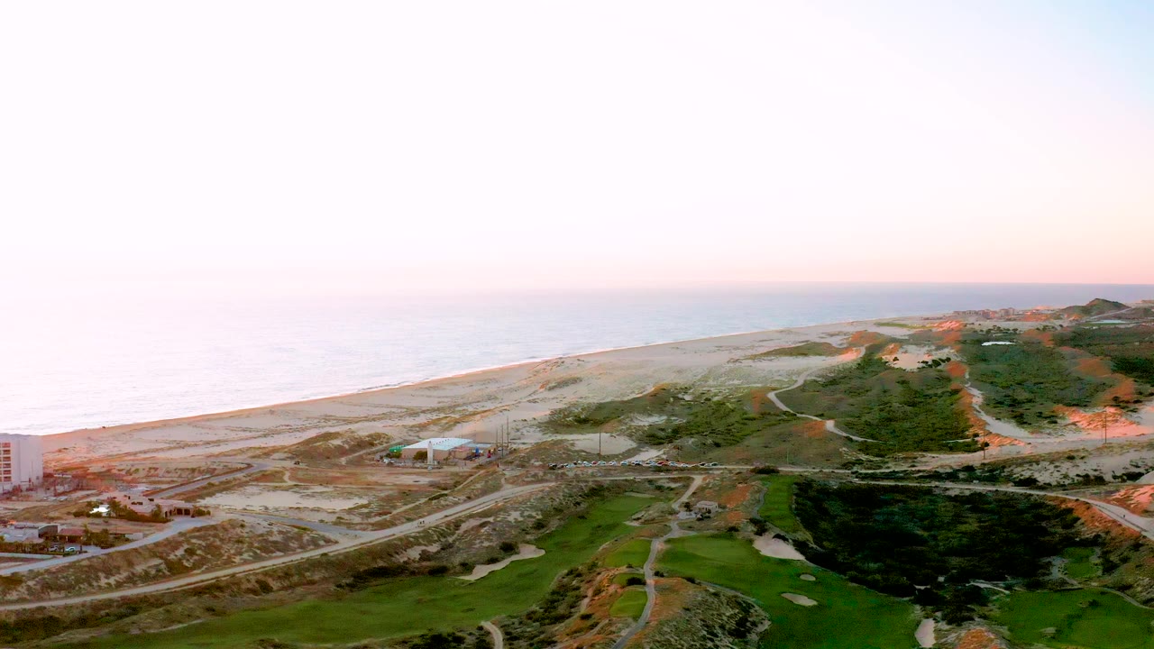 Diamante Cabo San Lucas coastline with a road running parallel to the beach, small vehicles on the road, green areas and hillside development to the right, and no visible people.
