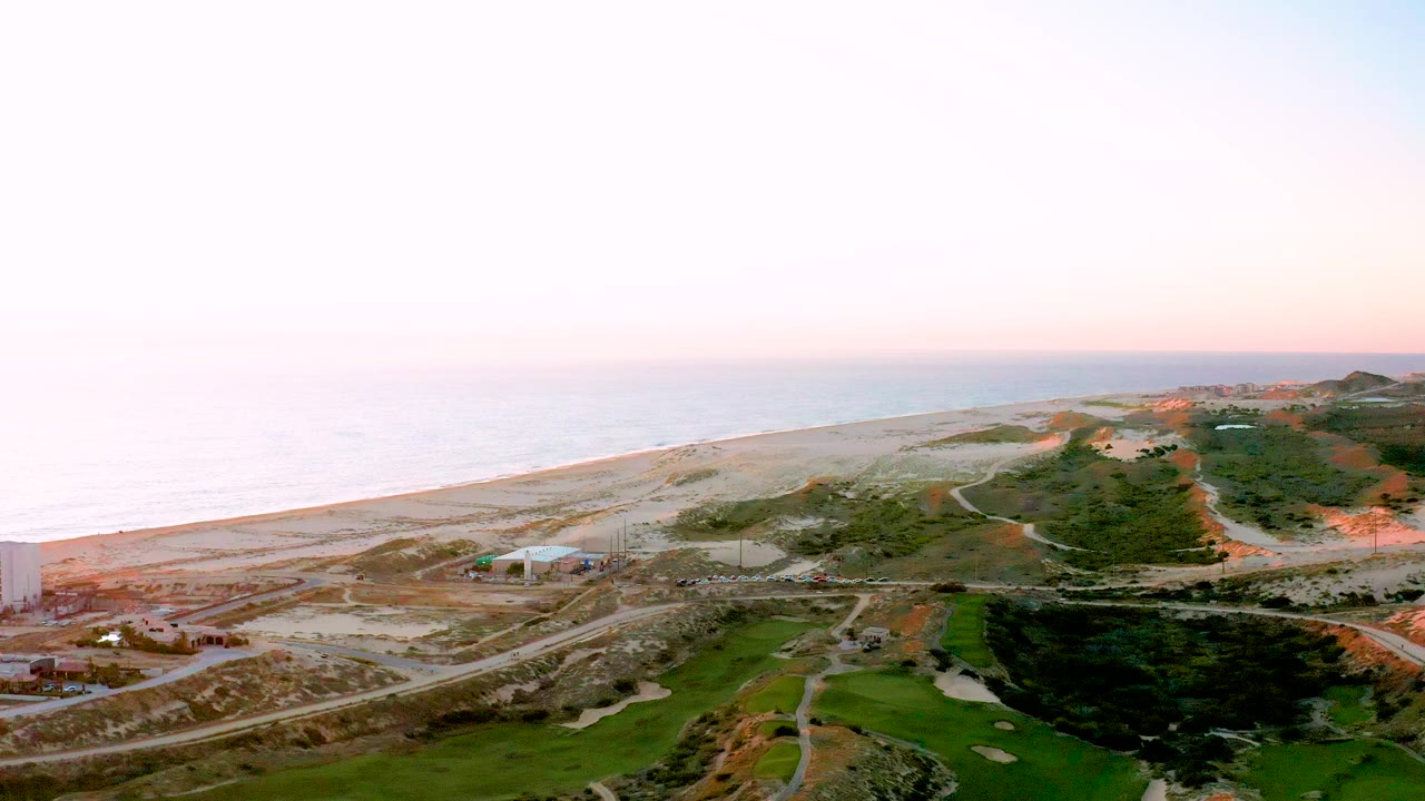 Aerial frame over Diamante Cabo San Lucas showing sandy coastline, ocean to the left, intersecting roads with vehicles, green grounds in the lower frame, and no visible people.