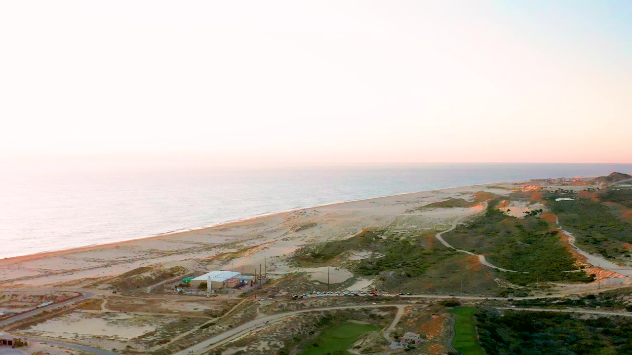Aerial frame over Diamante Cabo San Lucas showing pale beach and ocean surf to the left, scrubby dunes and dirt roads in the center, construction structures near the bottom, and no visible people.
