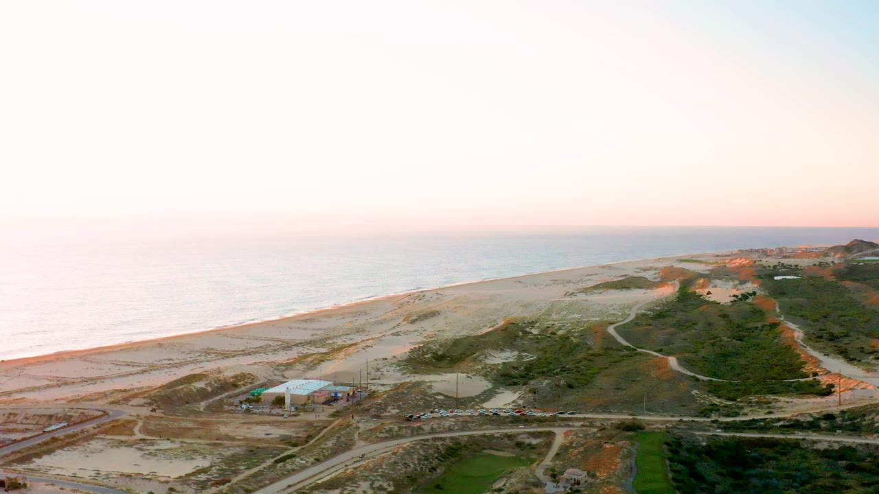 Drone-style frame over Diamante Cabo San Lucas with ocean haze on the left, beach and dunes crossing the view, small roads and construction structures below, low hills at the upper right, and no visible people.