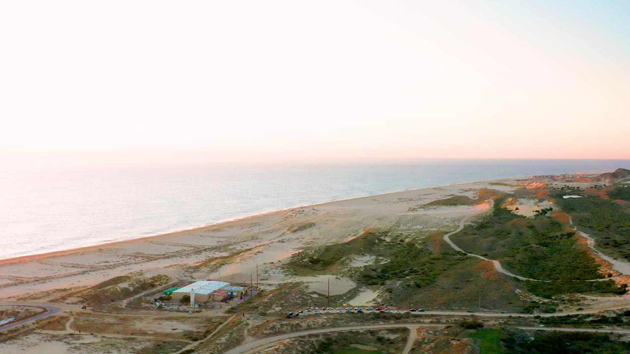 Opening aerial frame over Diamante Cabo San Lucas showing ocean at left, a long sandy beach, dune vegetation, dirt roads, a small light-roofed building, and no visible people.