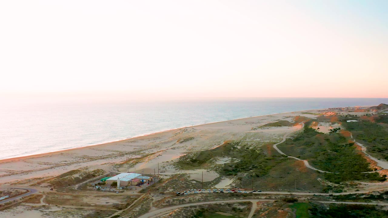 Second aerial frame of Diamante Cabo San Lucas with the pale ocean and beach on the left, dune paths through the center, the small building lower left, and no visible people.