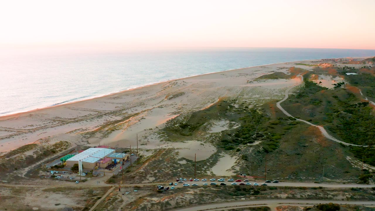 At 0:02, the aerial view in Diamante Cabo San Lucas shows the road and parked cars along the bottom edge, a small building with blue tarp, winding dune paths, ocean, and no visible people.