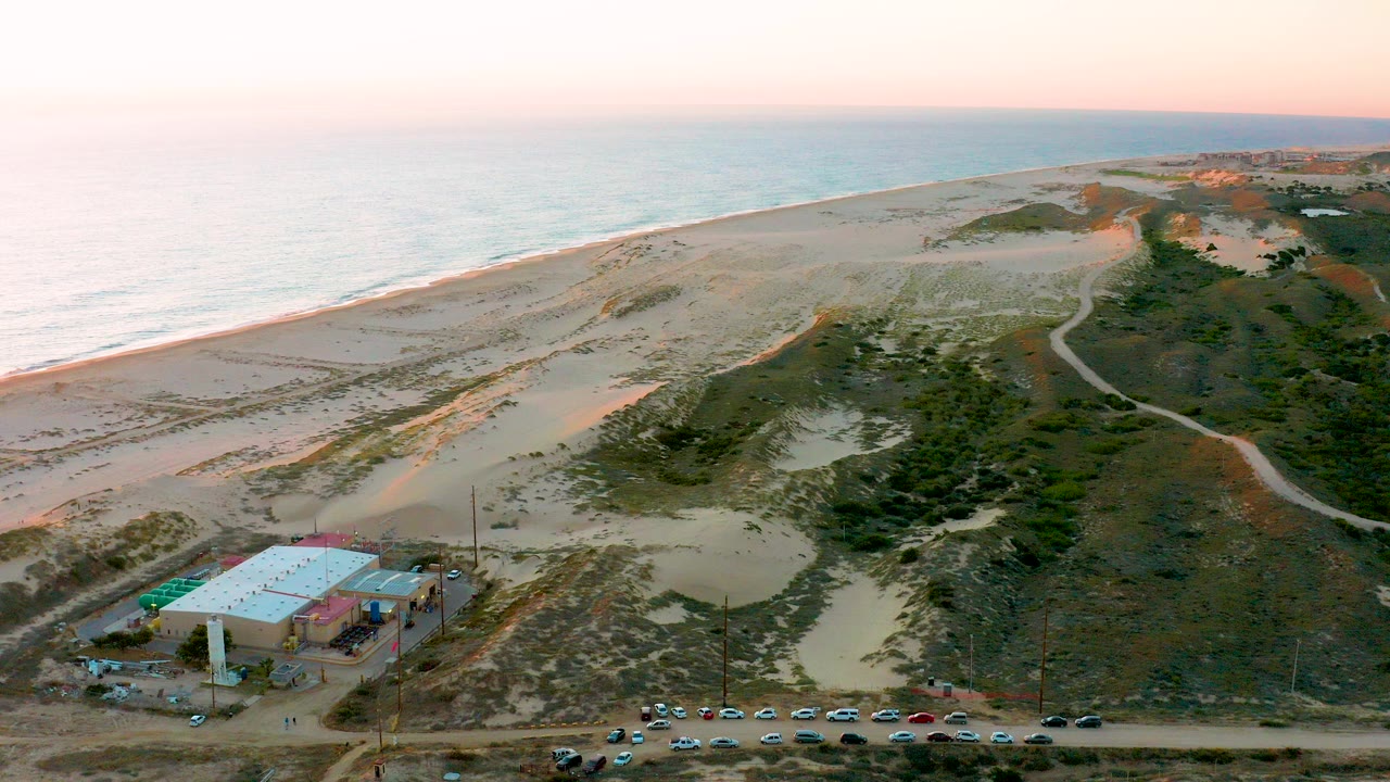Aerial view over Diamante Cabo San Lucas beach dunes with the ocean at left, a low building and parking lot near the bottom edge, and no visible people.