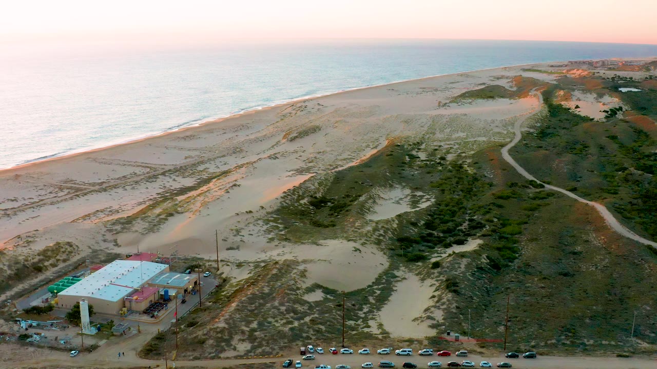 Aerial beach scene with dunes, scrub, ocean, road, and building near the lower left.