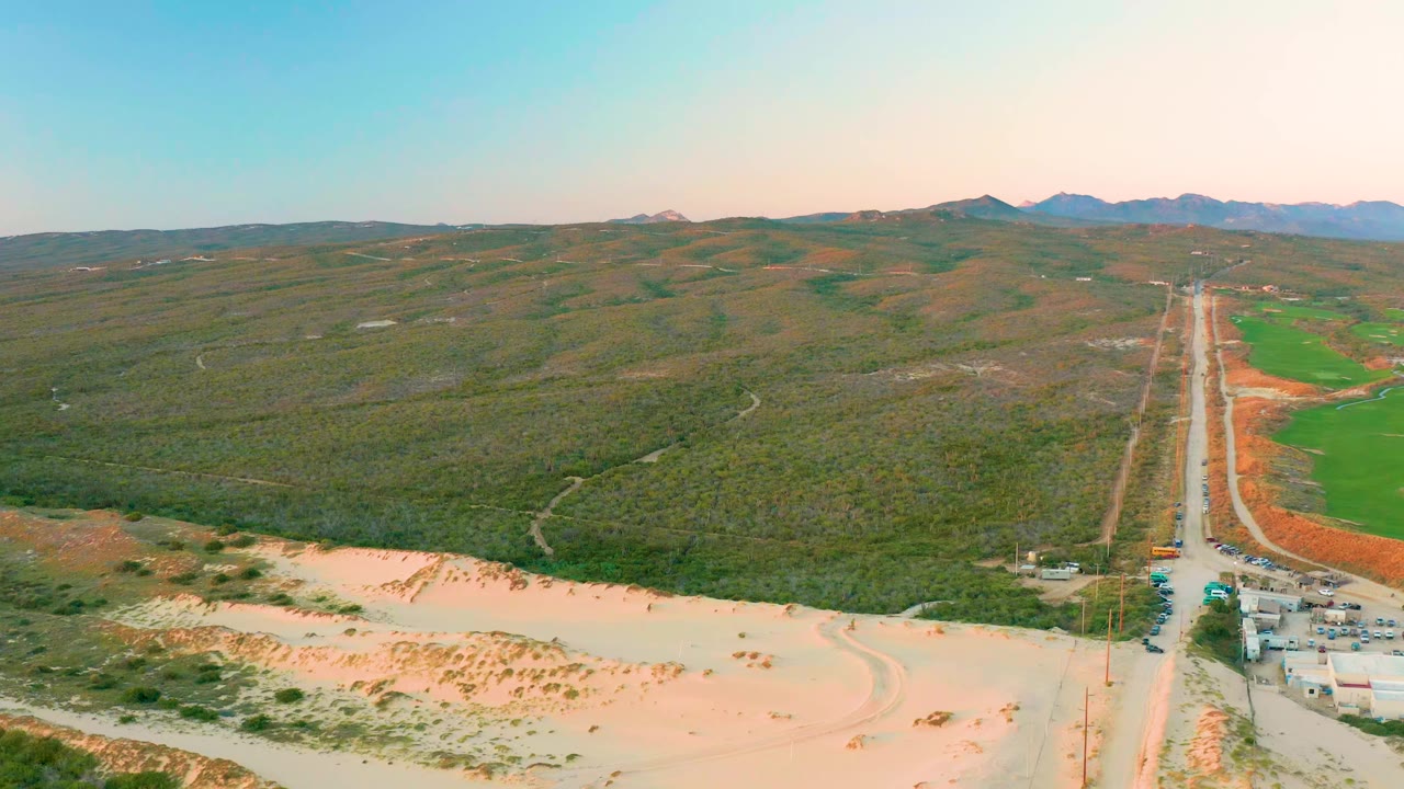 Elevated landscape with central road, sandy foreground, green fields, and hills.