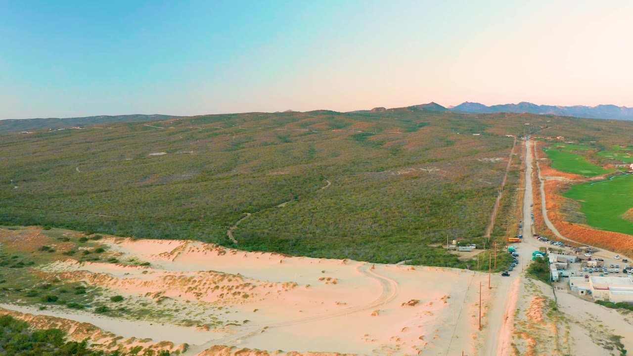 High aerial view in Diamante Cabo San Lucas with the road centered slightly right, sandy ground in front, hilly brushland across the middle, green fields to the right, and no visible people.