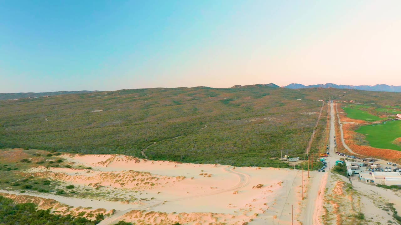 Aerial view in Diamante Cabo San Lucas with the road centered to the right, vehicles clustered near the roadside, pale sand in front, green hills beyond, and no visible people.