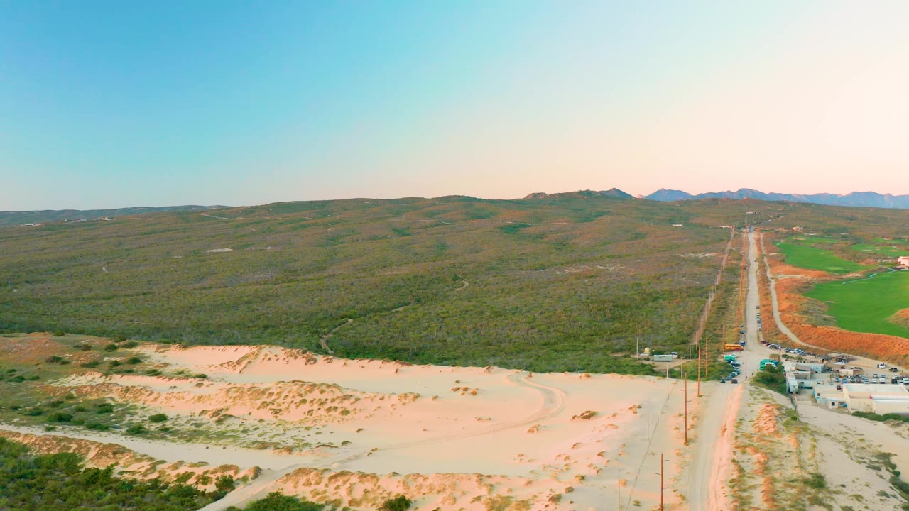High aerial frame over Diamante Cabo San Lucas showing a long road receding toward hills, sandy ground across the foreground, green cultivated fields on the right, small vehicles, and no visible people.
