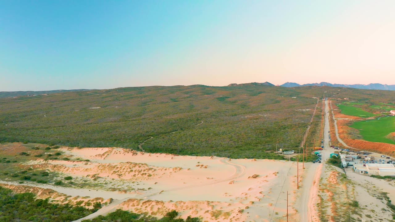 Aerial frame in Diamante Cabo San Lucas with pale sandy terrain at bottom, a rural road and utility poles on the right, vehicles near a small roadside area, green hills, and no visible people.