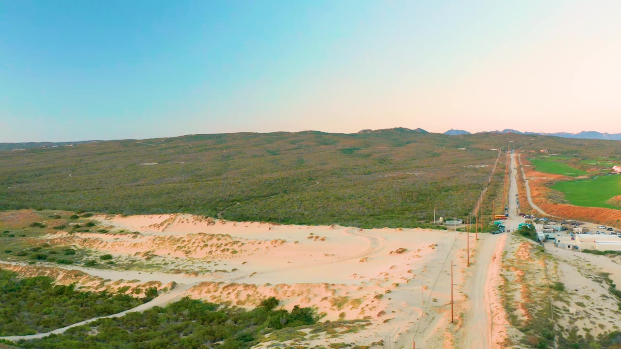 Drone view in Diamante Cabo San Lucas with sandy foreground, scrub vegetation, a straight road, distant low hills, green fields at right, and no visible people.