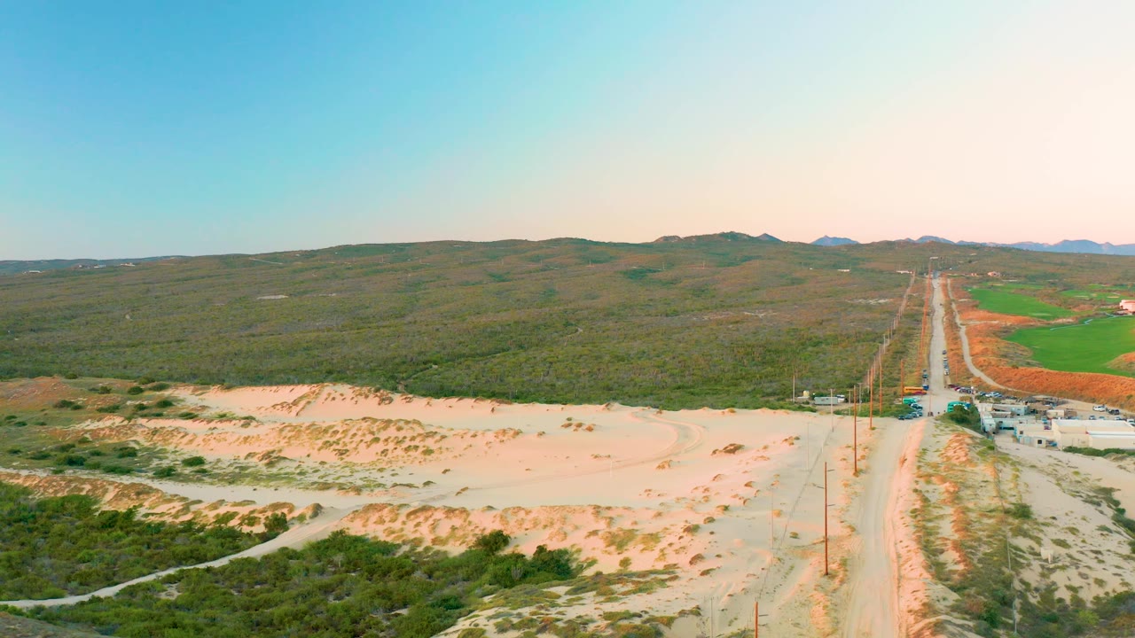 High aerial frame in Diamante Cabo San Lucas with dunes across the foreground, a rural road at right, scrubland stretching to hills, green fields, and no visible people.