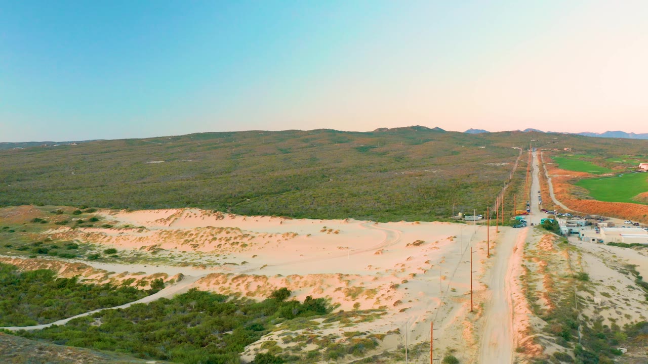 Aerial landscape with sandy foreground, road, utility poles, and low hills.
