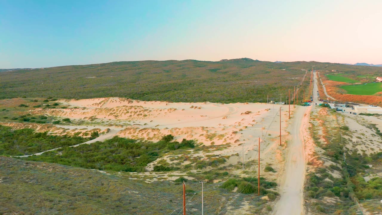 Aerial view in Diamante Cabo San Lucas with the dirt road slightly right of center, power poles along both sides, pale sandy dunes, green brush, distant hills, and no visible people.