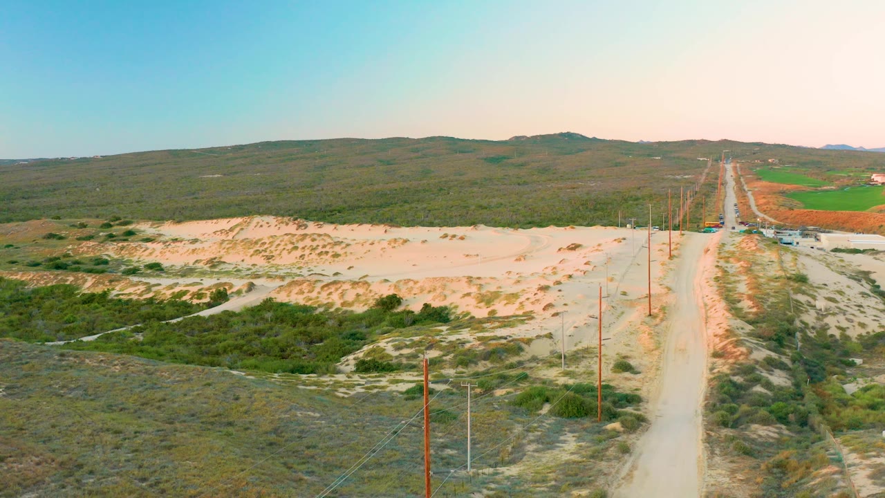 Aerial view in Diamante Cabo San Lucas with sandy terrain filling the left side, a straight dirt road and pole line on the right, broad scrubland below, low hills beyond, and no visible people.