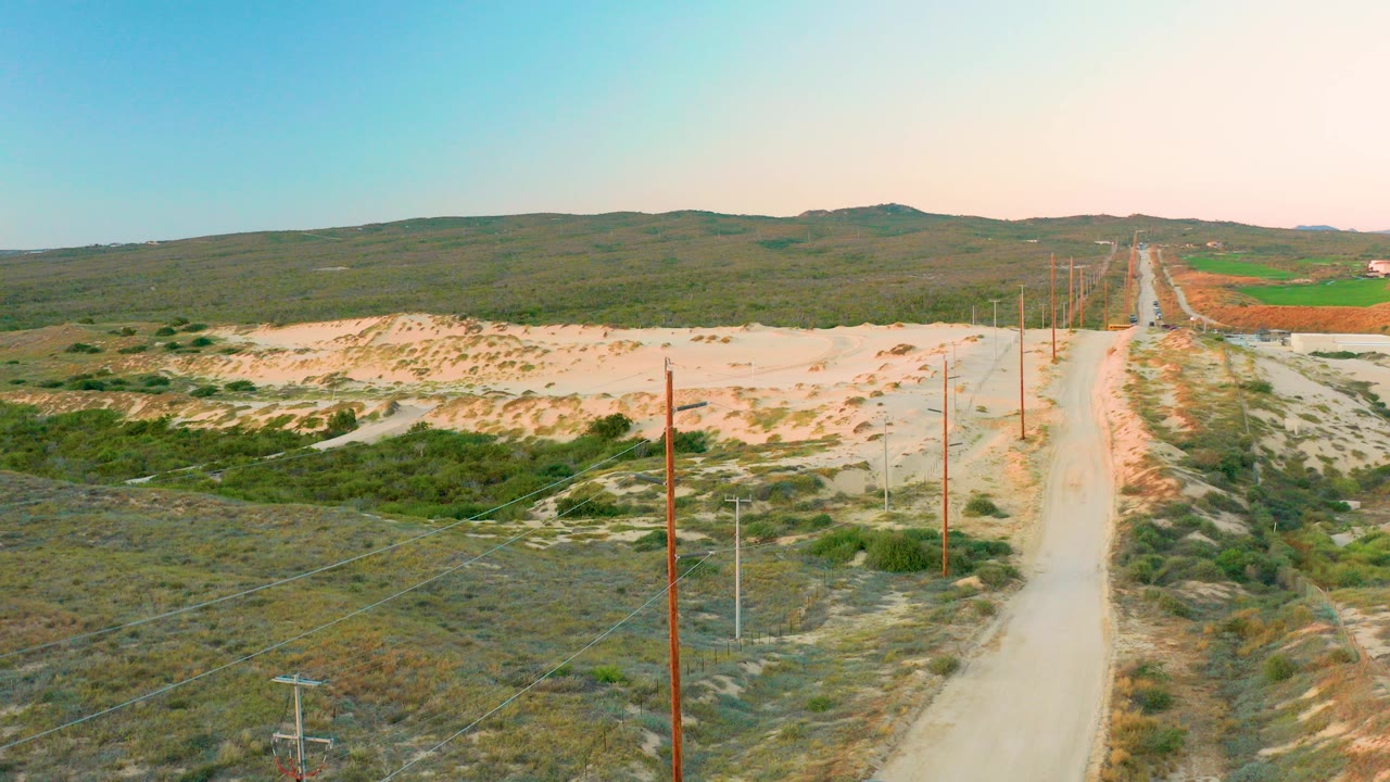 Aerial view over Diamante Cabo San Lucas showing a pale dirt road beside utility poles, dry scrub vegetation, sandy ground, green field patches, distant hills, and no visible people.