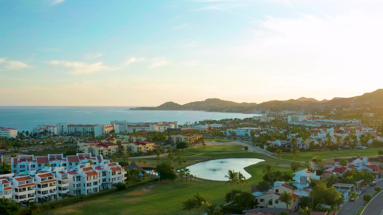 Wide aerial frame over San Jose del Cabo showing white residential buildings, a green golf course, a reflective water hazard, ocean, mountains, warm sky, and no visible people.