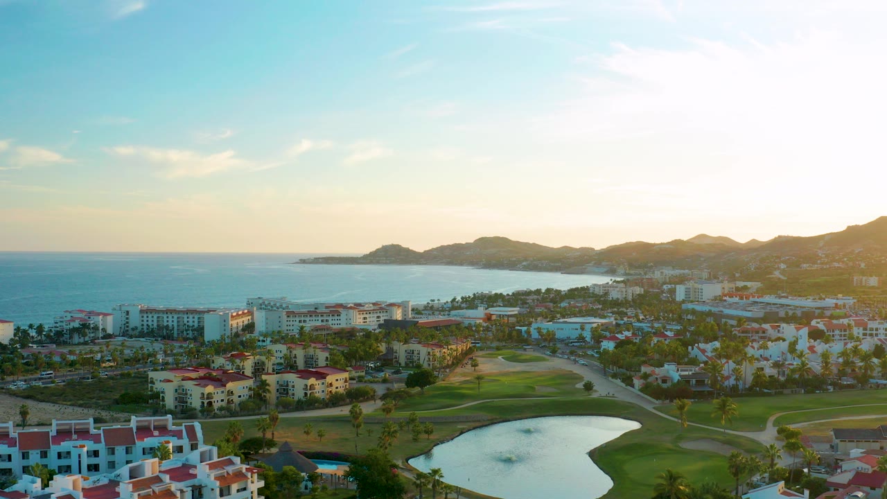 Opening aerial frame over San Jose Del Cabo with a golf course pond in the foreground, resort buildings and palms in the midground, ocean and low mountains beyond, and no visible people.