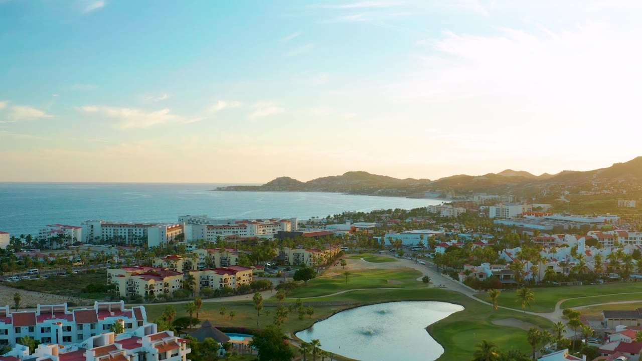 Coastal resort aerial with golf course, pond, palms, ocean, and hills.