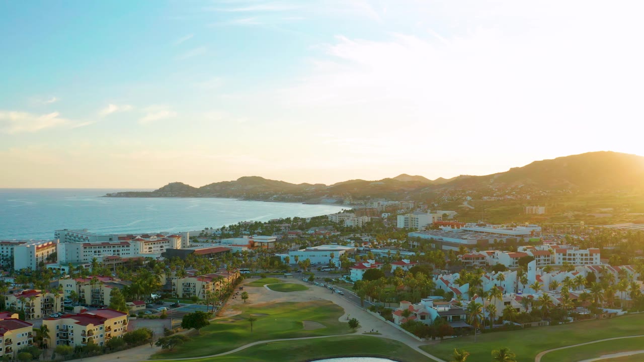 San Jose del Cabo aerial view with the bay stretching along the left edge, resort buildings below, green fairway in front, layered hills beyond the shoreline, and no visible people.