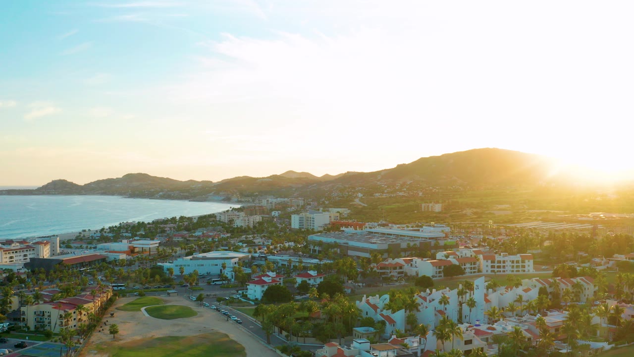 Opening wide aerial view of San Jose del Cabo with ocean on the left, low mountains on the horizon, golf course and buildings below, bright sun flare at right, and no visible people.