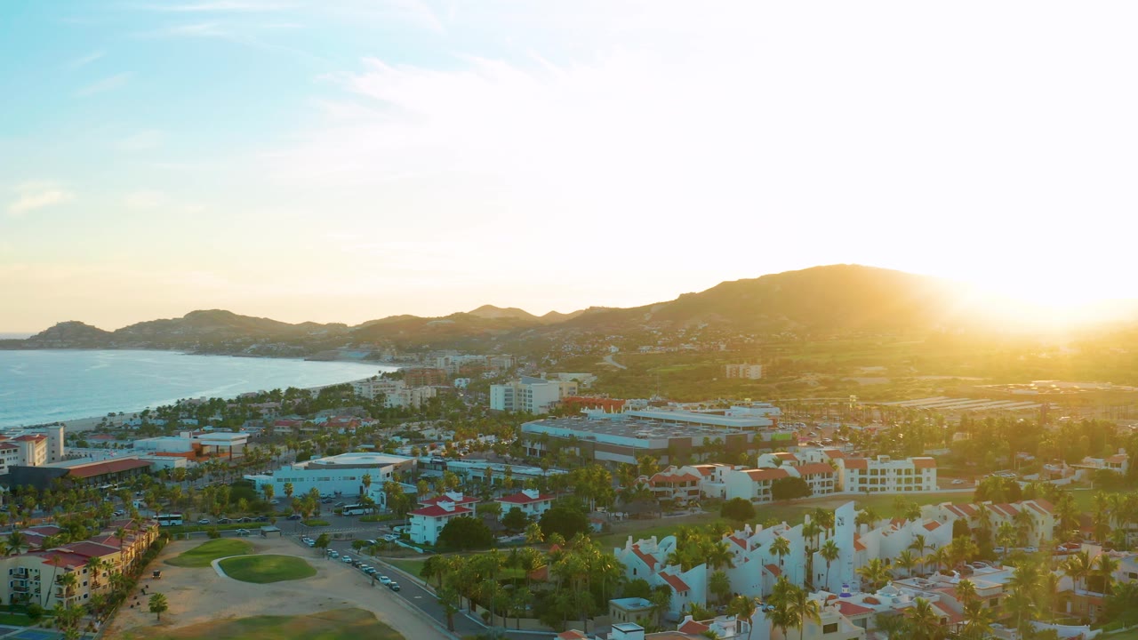 Wide San Jose del Cabo sunset frame with palms and low buildings in the foreground, a golf course at lower left, ocean along the left edge, mountains across the background, and no visible people.
