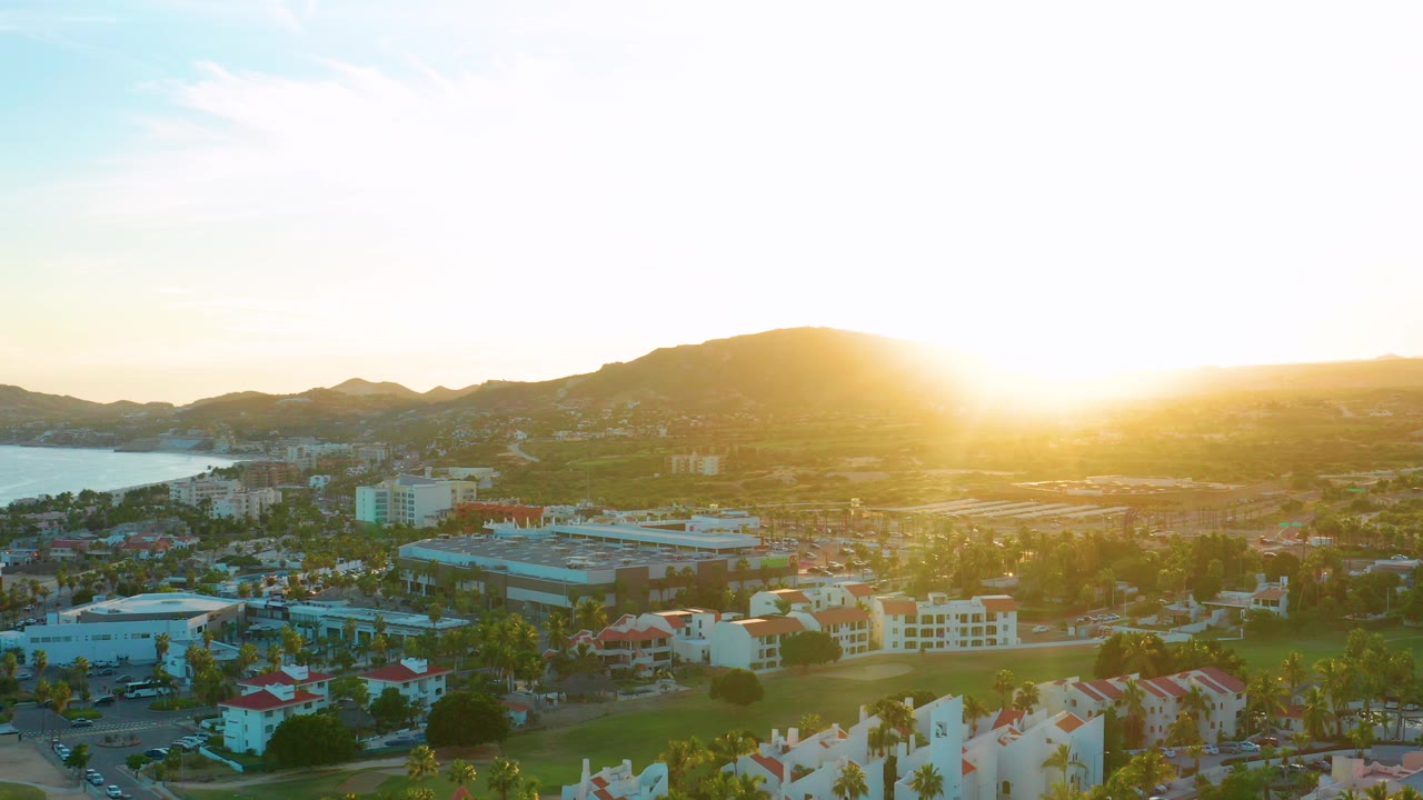 San Jose del Cabo aerial view at sunset with water on the left, low buildings and palms below, mountain ridges on the horizon, and no visible people.