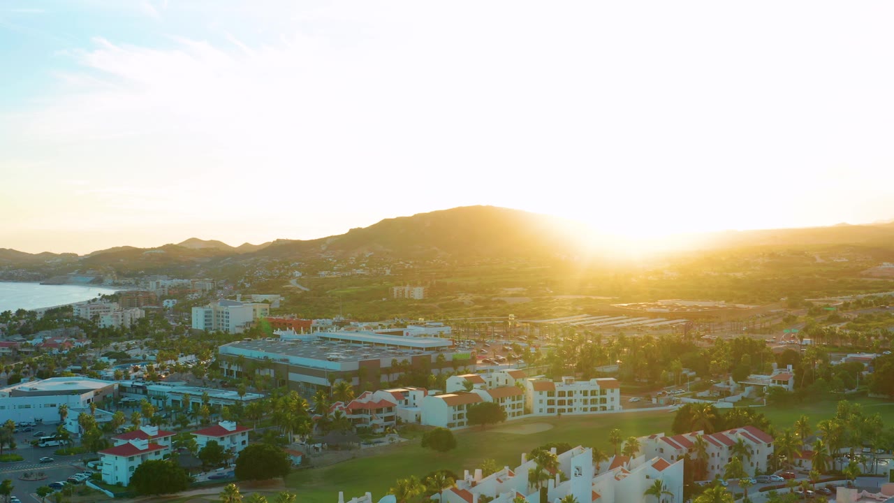 San Jose del Cabo is seen from above with red and white roofs in the lower frame, a strip of water at far left, hazy mountain silhouettes, and no visible people.