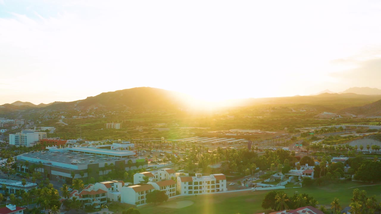 Aerial view over San Jose Del Cabo with the sun low above a mountain ridge, city buildings and trees below, and no visible people.