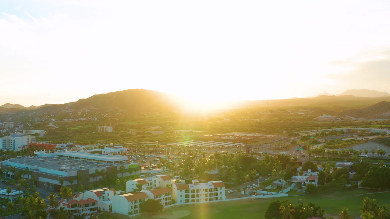 A high view of San Jose Del Cabo shows backlit rooftops, greenery, roads, and a mountain horizon under intense sunset glare with no visible people.