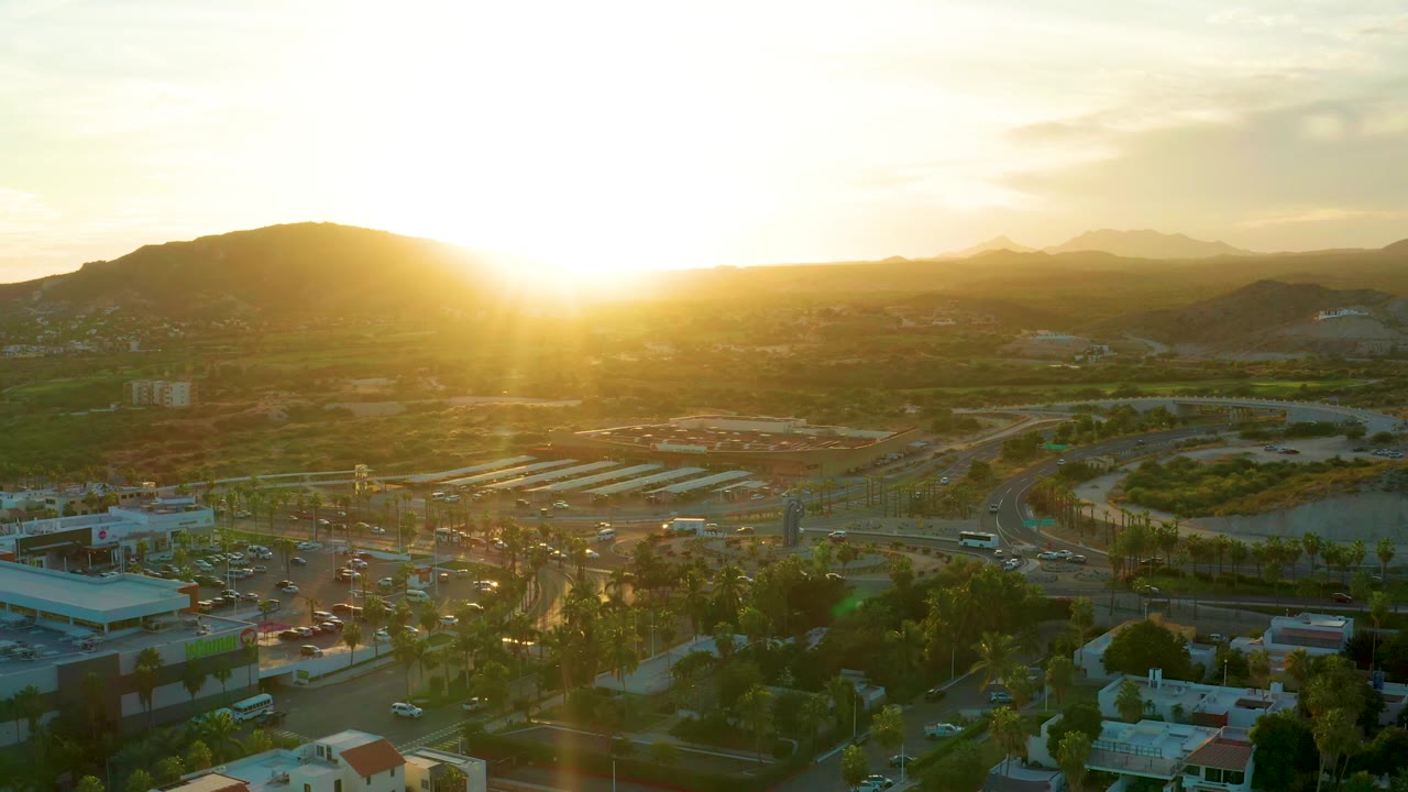 San Jose del Cabo aerial frame with golden sun glare over a curving road system, low buildings, trees, distant hills, and no visible people.